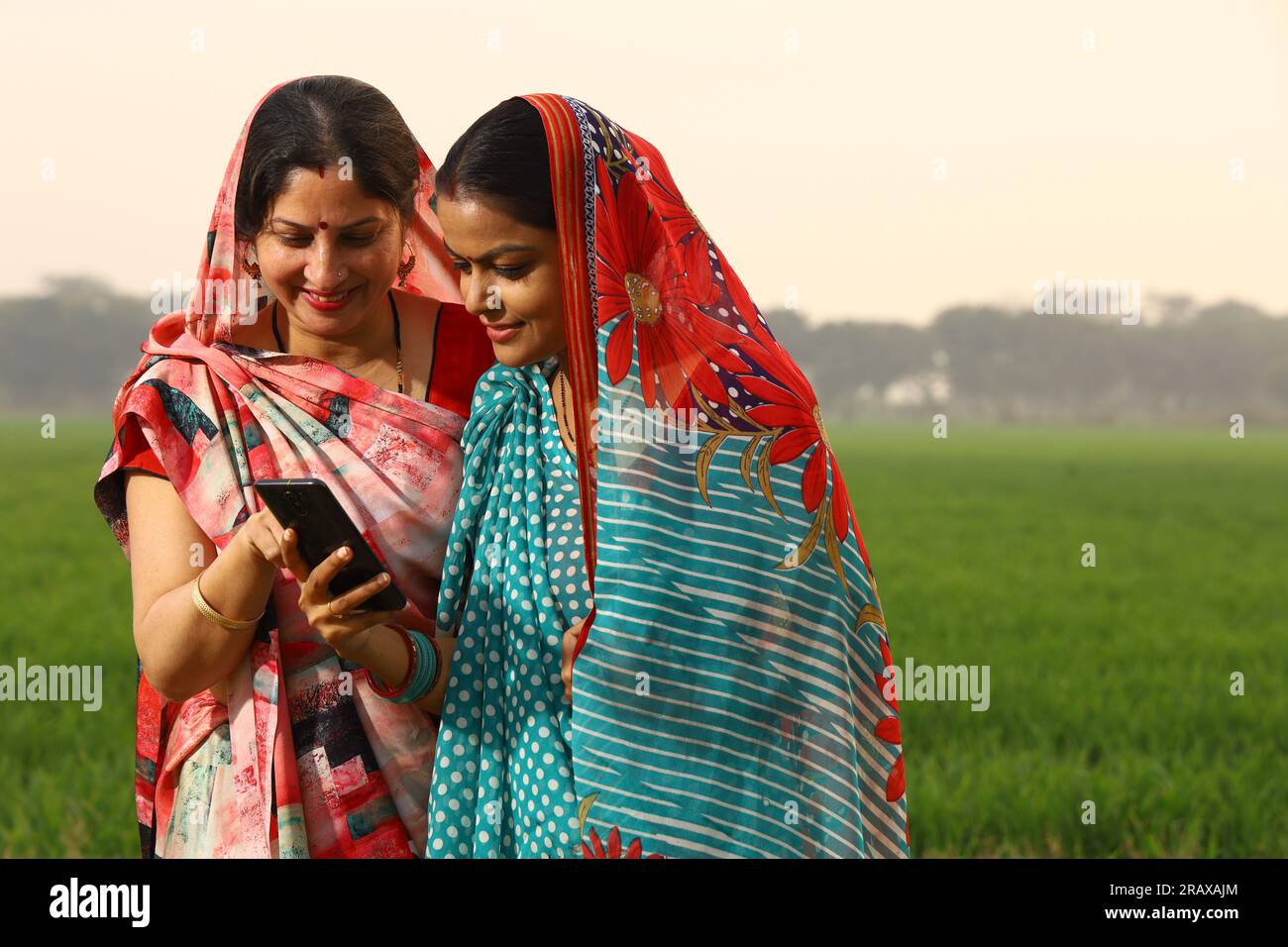Glückliche, ländliche indische Frauen, die auf einem Senf-Ackerfeld stehen und mit dem Handy in der Hand surfen. Stockfoto
