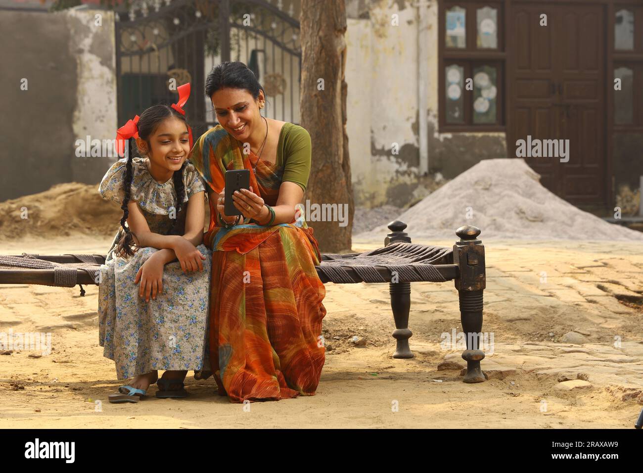 Glückliche indische Landfamilie im Dorf. Mutter und Tochter sitzen zusammen und lächeln auf dem Kinderbett vor ihrem Haus mit dem Handy. Stockfoto