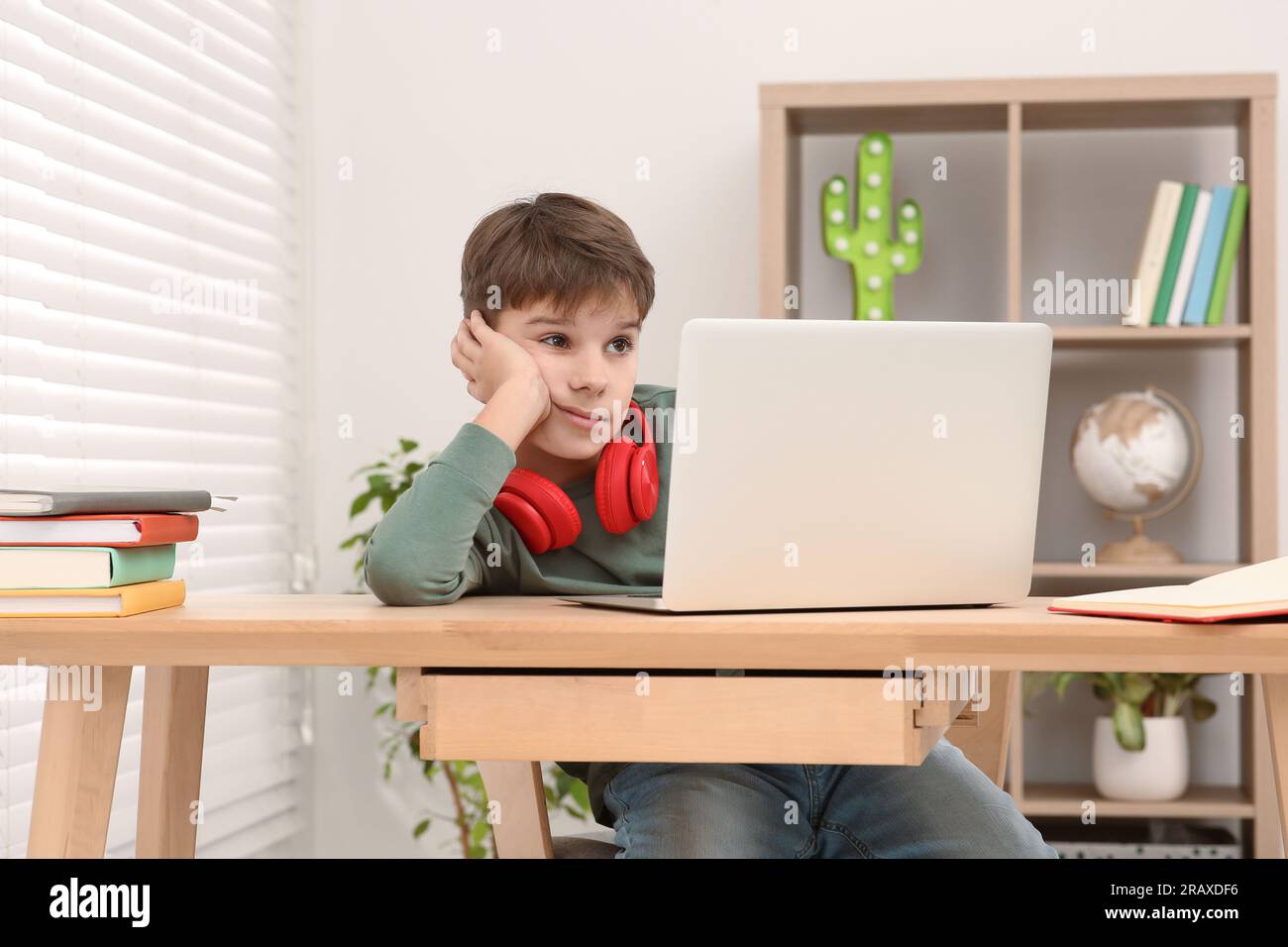 Müder Junge mit roten Kopfhörern, der am Schreibtisch im Zimmer ein Notebook benutzt. Zu Hause Stockfoto