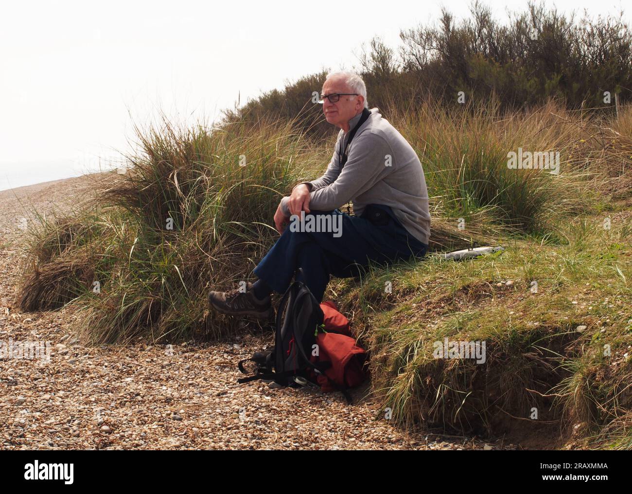 Ein Mann über 60 sitzt hinten am Kieselstrand in Minsmere, Suffolk in der Sonne, inmitten des Marram Grases Stockfoto