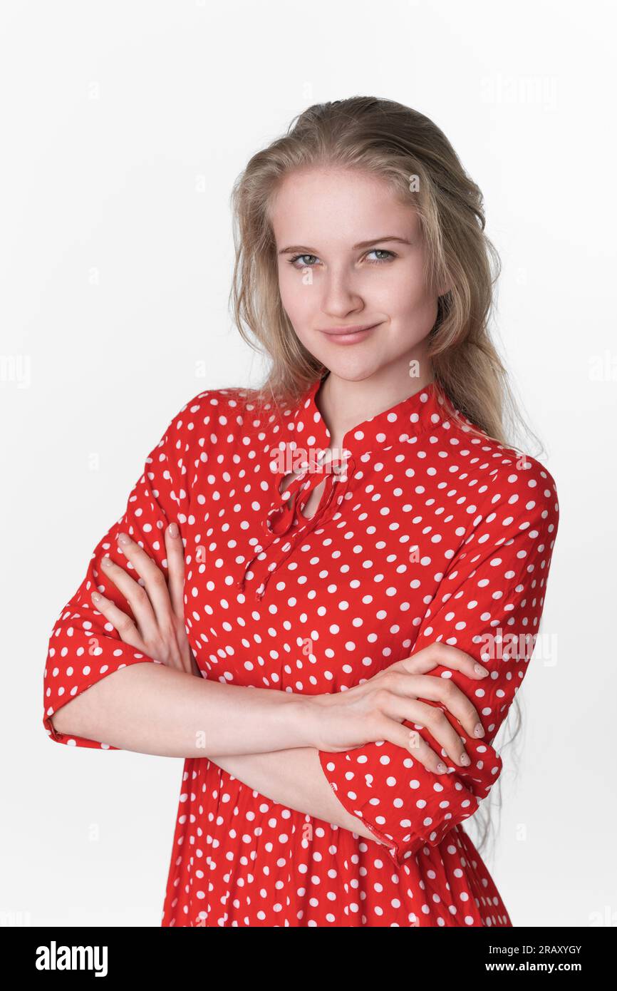 Porträt einer aufrichtig lächelnden jungen Frau, die Arme über die Brust gekreuzt und in die Kamera schaut. Charmantes europäisches weibliches Model, 21 Jahre alt, in rotem Kleid Stockfoto