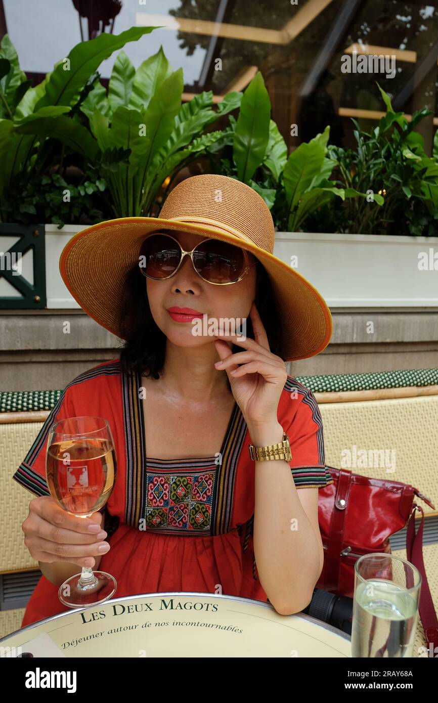 Sommer im Les Deux Magots, eine elegant gekleidete Frau mit Glas, die an einem Café-Tisch auf der Außenterrasse am linken Ufer von Paris sitzt. Stockfoto