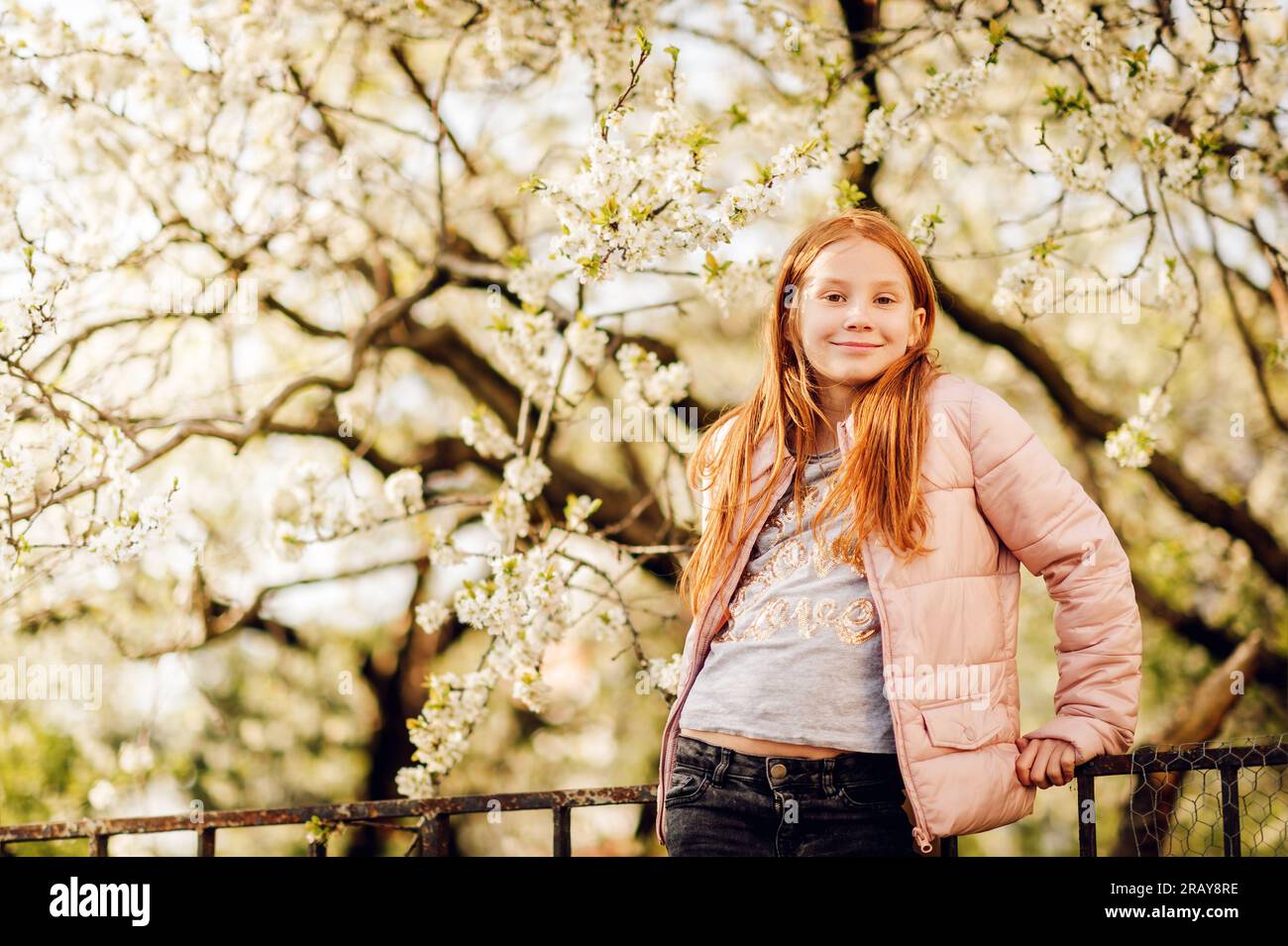 Fröhliches Mädchen, das draußen mit blühendem Apfelbaum spielt und eine pinkfarbene wattierte Jacke trägt Stockfoto