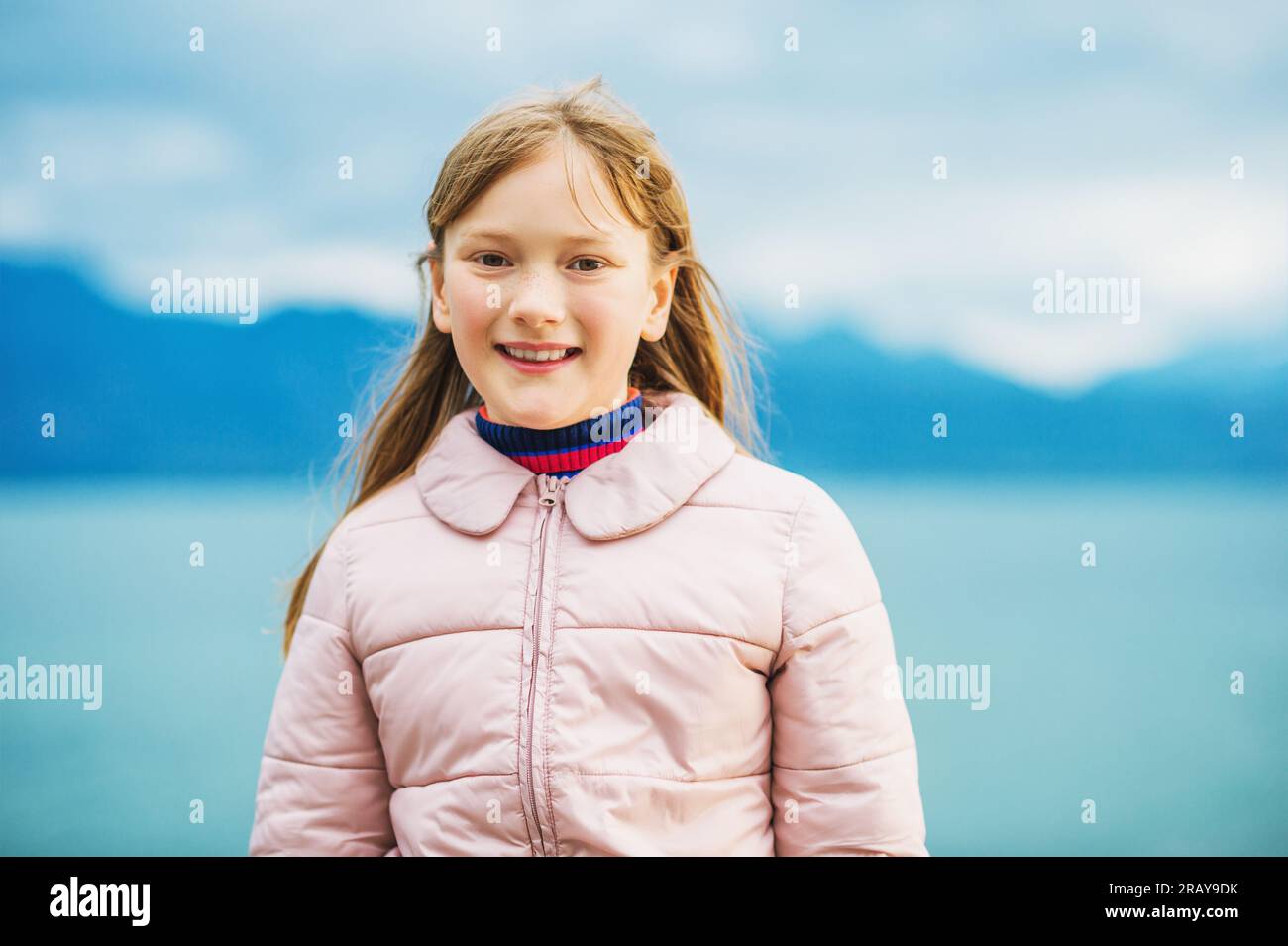 Süßes kleines 9-10-jähriges Mädchen, das an einem sehr windigen Tag am See spielt und eine warme rosa Jacke trägt Stockfoto