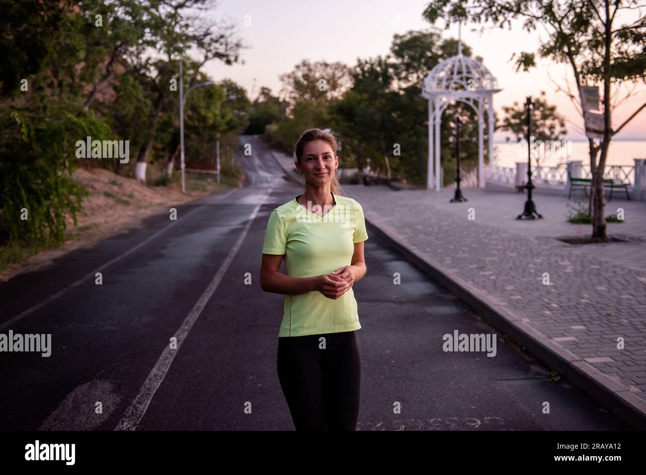 Eine junge Frau in Sportbekleidung läuft im Morgengrauen den Weg entlang. Morgenroutine, Meditation, Gefühl der Freiheit. Das Mädchen trägt schwarze Leggings und ein gelbes T-Shirt. Stockfoto