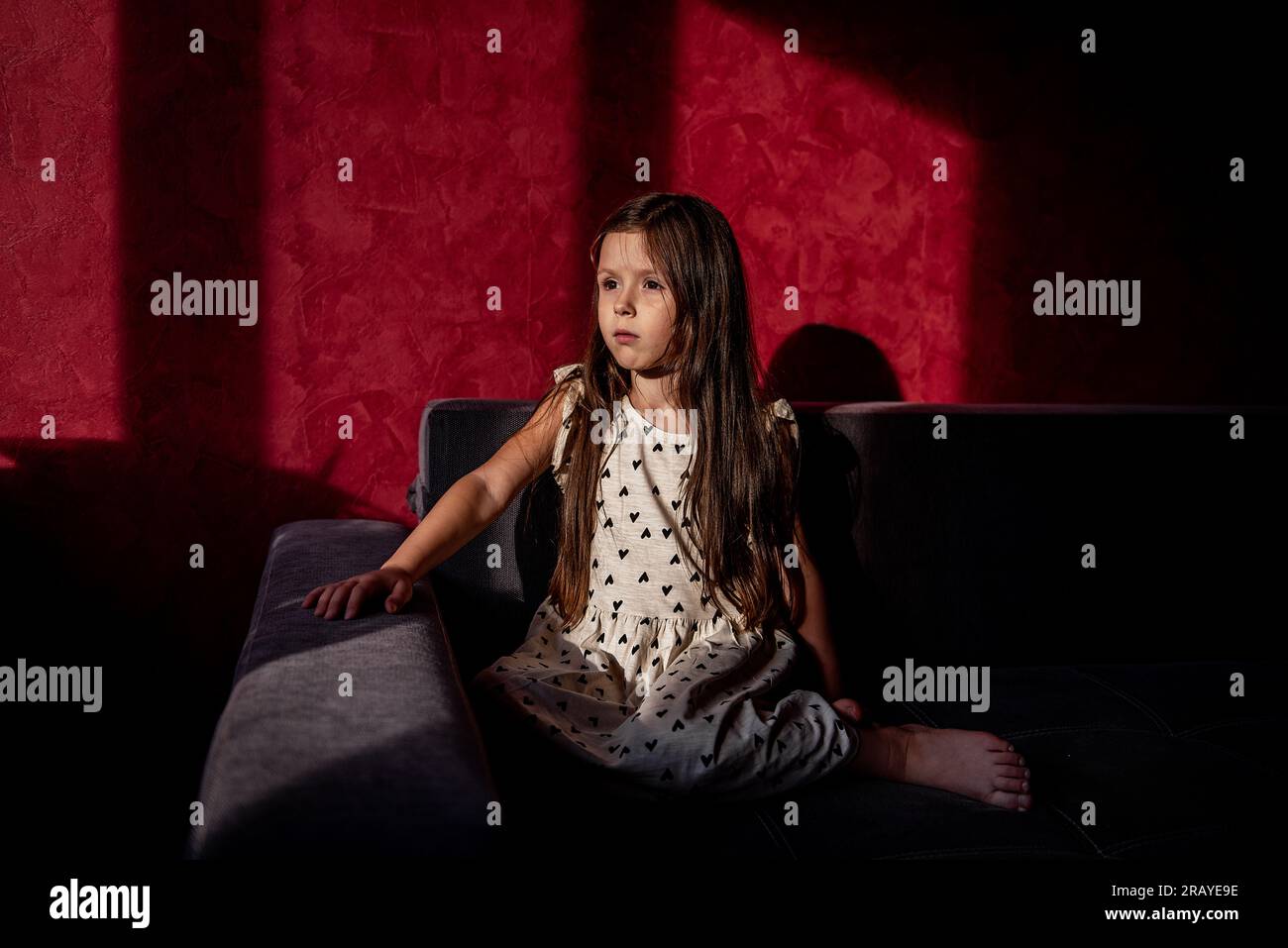 Das Porträt eines kleinen Mädchens mit langen Haaren sitzt auf dem Sofa vor rotem Hintergrund mit hellem Licht und Schatten aus dem Fenster. Das Mädchen schaut mit in die Kamera Stockfoto