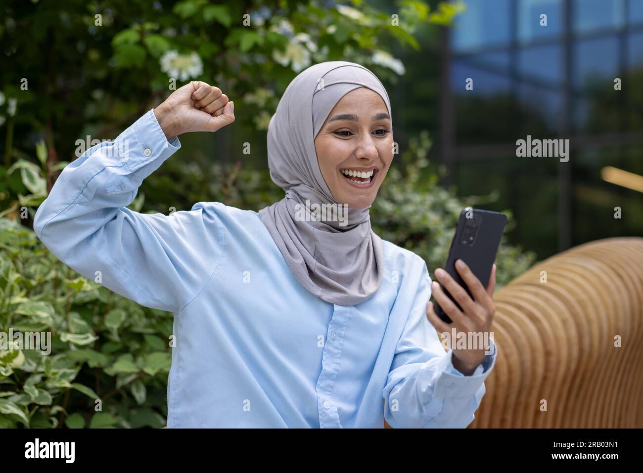 Die erfolgreiche, fröhliche muslimische Frau in Hijab erhielt eine Online-Gewinnbenachrichtigung, Dinka las Nachrichten und hielt die Hand hoch, um die Triumph-Geste zu feiern, als sie auf einer Bank vor dem Bürogebäude im Park saß. Stockfoto