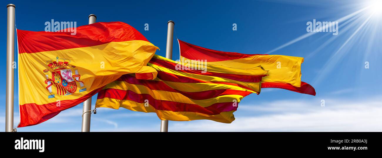 Nahaufnahme der spanischen und katalanischen Flaggen (La Rojigualda und Senyera) mit Fahnenmast, der auf einem blauen Himmel mit Wolken, Sonnenstrahlen und Kopierraum im Wind weht Stockfoto