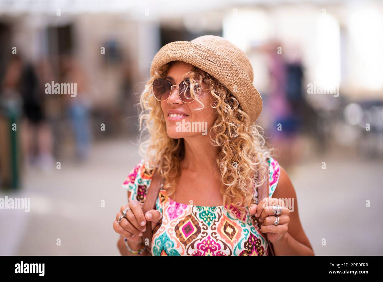 Glückliche Touristin mit blonden langen lockigen Haaren und Hut, die sich umsieht und alleine mit dem Rucksack läuft, Freizeitaktivitäten und Urlaubszeit genießt. Menschen im Hintergrund unscharf gemacht. Stockfoto