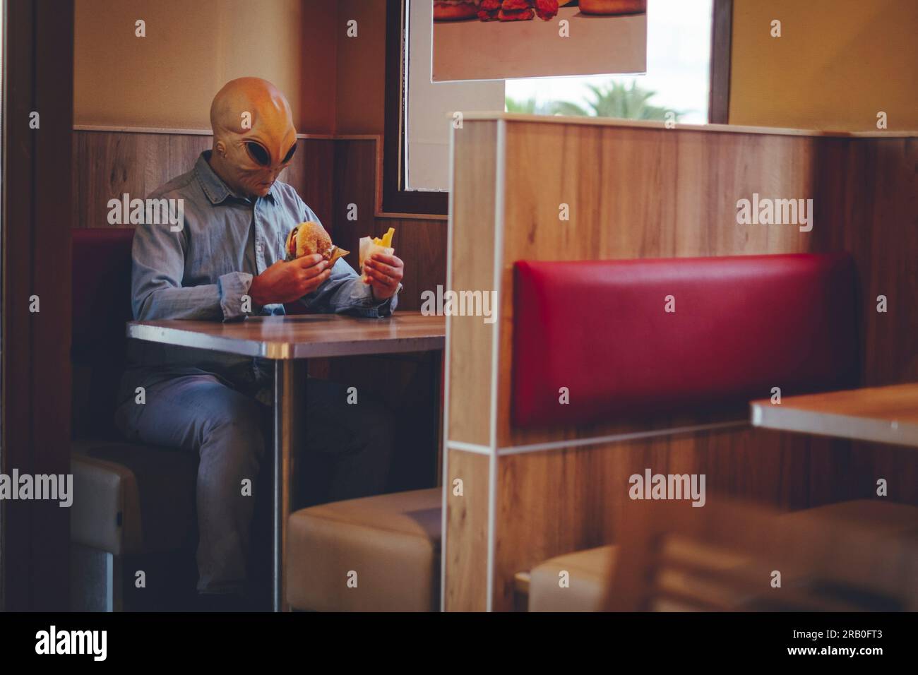 Ein Mann mit Alien-Maske, der allein in einem Fast-Food-Laden isst, schlechte Hamburger und pommes frites. UFO leben unter uns als menschliches Konzept, komisches Bild. Invasion und Diversität Menschen in Freizeitzimmern Stockfoto