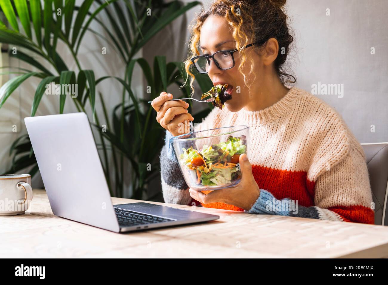 Eine Frau, die mit Laptop beschäftigt ist und gleichzeitig Salate isst. Kurze Mittagspause für Online-Aktivitäten. Diätkonzept und moderne Arbeit. Zwanglose Frau in Telearbeit allein im intelligenten Arbeitsleben Stockfoto