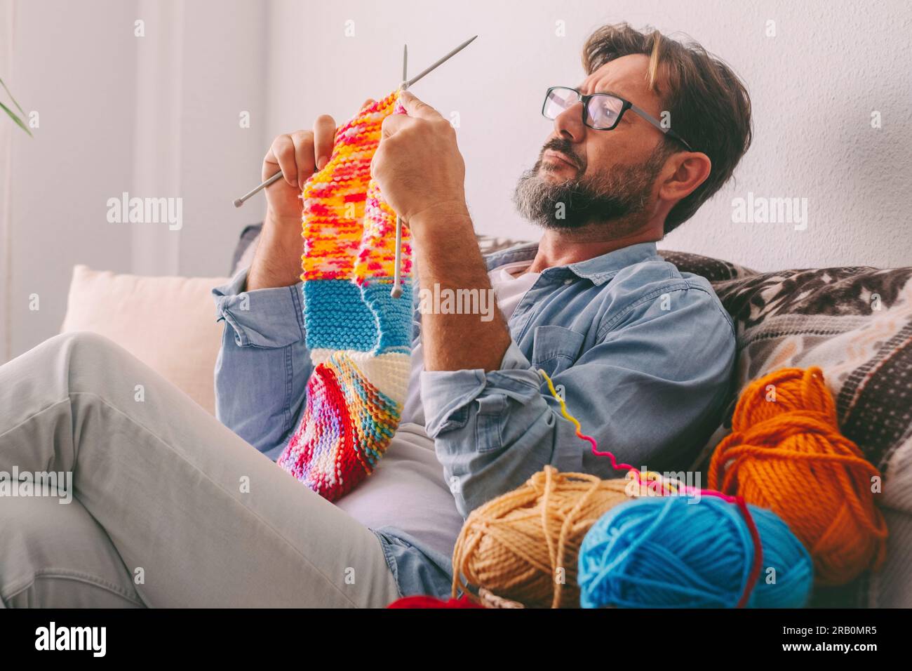Ein junger, reifer Mann strickt zu Hause mit Wolle und Nadeln und sitzt bequem auf dem Sofa bei der üblichen weiblichen Freizeitbeschäftigung im Innenbereich. Diversität und Guitality weiblich männlich. Leute, die drinnen arbeiten Stockfoto