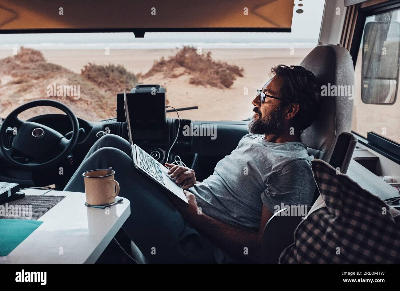Reisende genießen Entspannung bei Freizeitaktivitäten im Haus, arbeiten und verwenden Laptop, sitzen auf dem Beifahrersitz in einem Wohnmobil vor einem Strand. Digitaler Nomaden-Lebensstil für Touristen Stockfoto