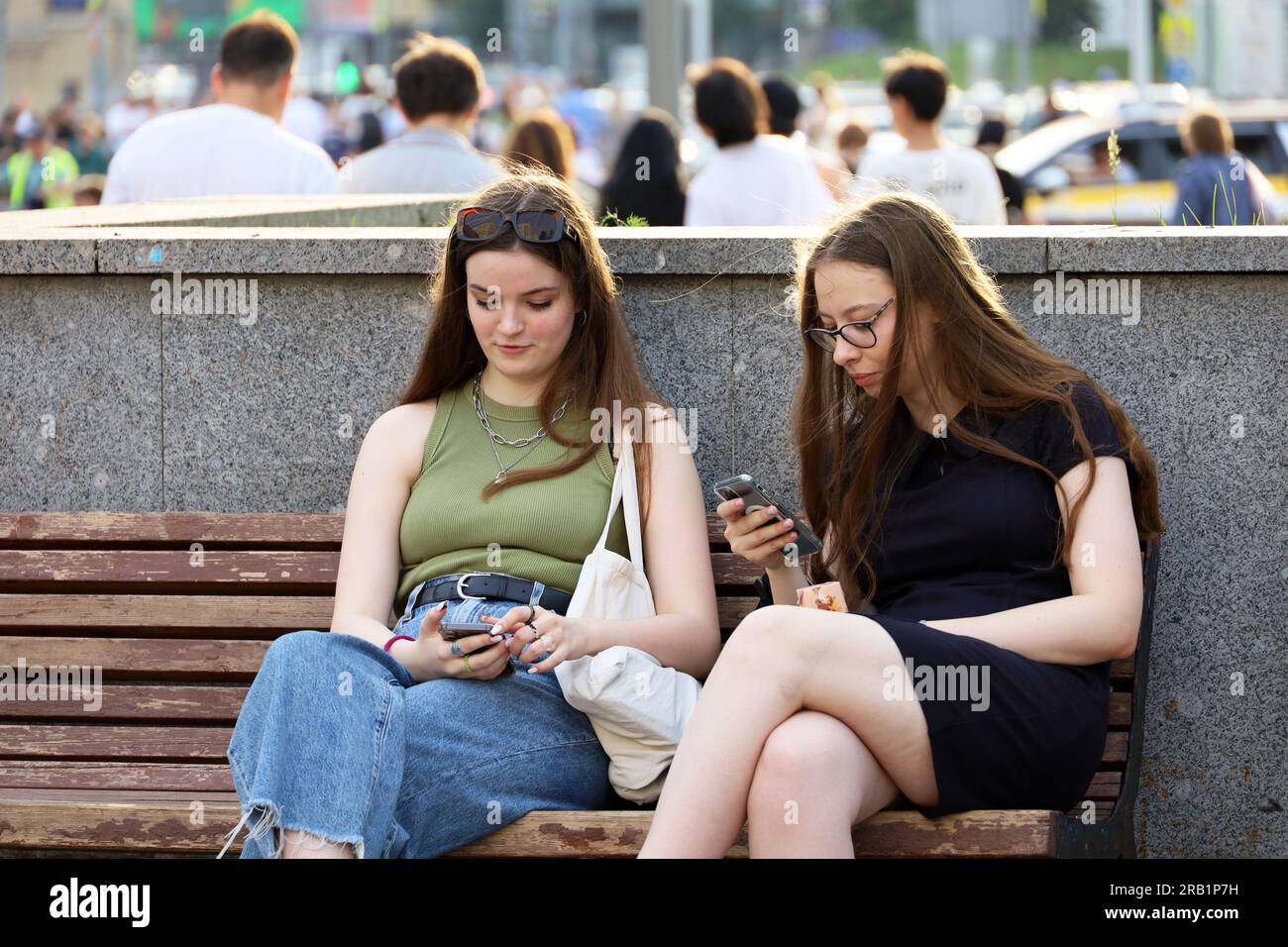 Zwei hübsche Mädchen, die auf einer Straße sitzen und Smartphones benutzen. Freundinnen mit Handys in der Sommerstadt Stockfoto