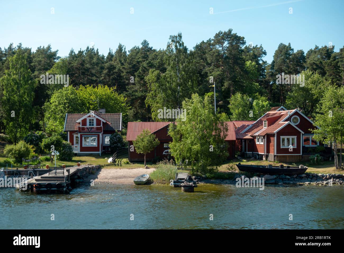 Typisches schwedisches rotes Haus mit weißen Verzierungen am Ufer in idyllischer Sommerlandschaft am Ufer mit privatem Anlegesteg und Boot für Urlaub Konzept Stockfoto