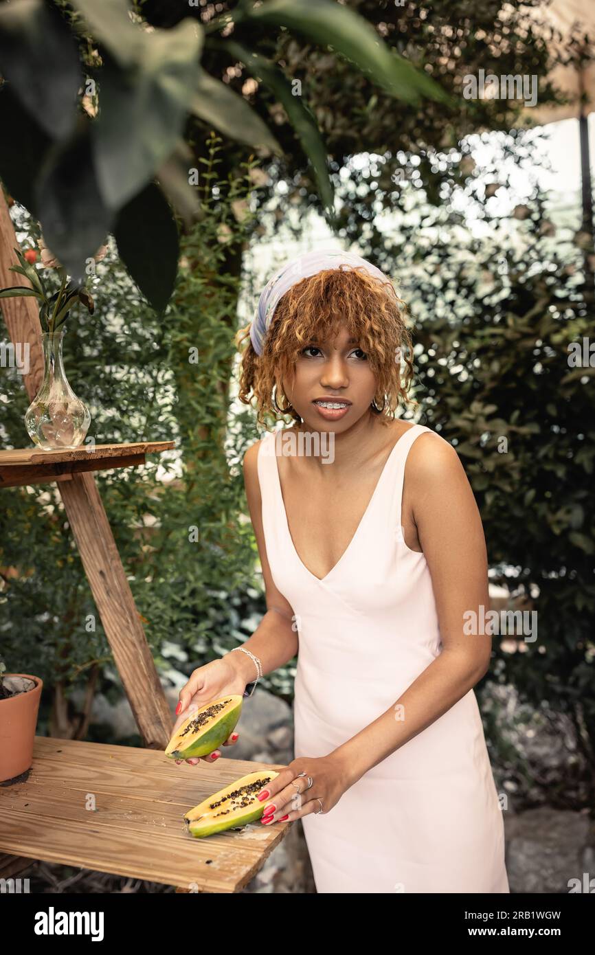 Lächelnde junge afroamerikanische Frau im Kopftuch und Sommerkleid, die geschnittene und reife Papaya hielt, während sie neben grünen Pflanzen im Grünen stand, Lady ins Stockfoto