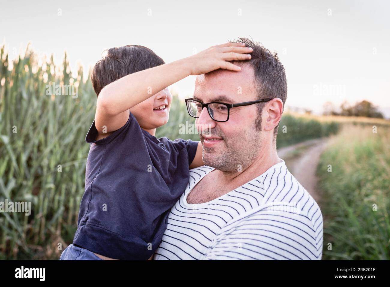 Eine Tochter mit dunklen kurzen Haaren spielt mit der Brille ihres Vaters, der sie im Maisfeld in den Händen hält Stockfoto