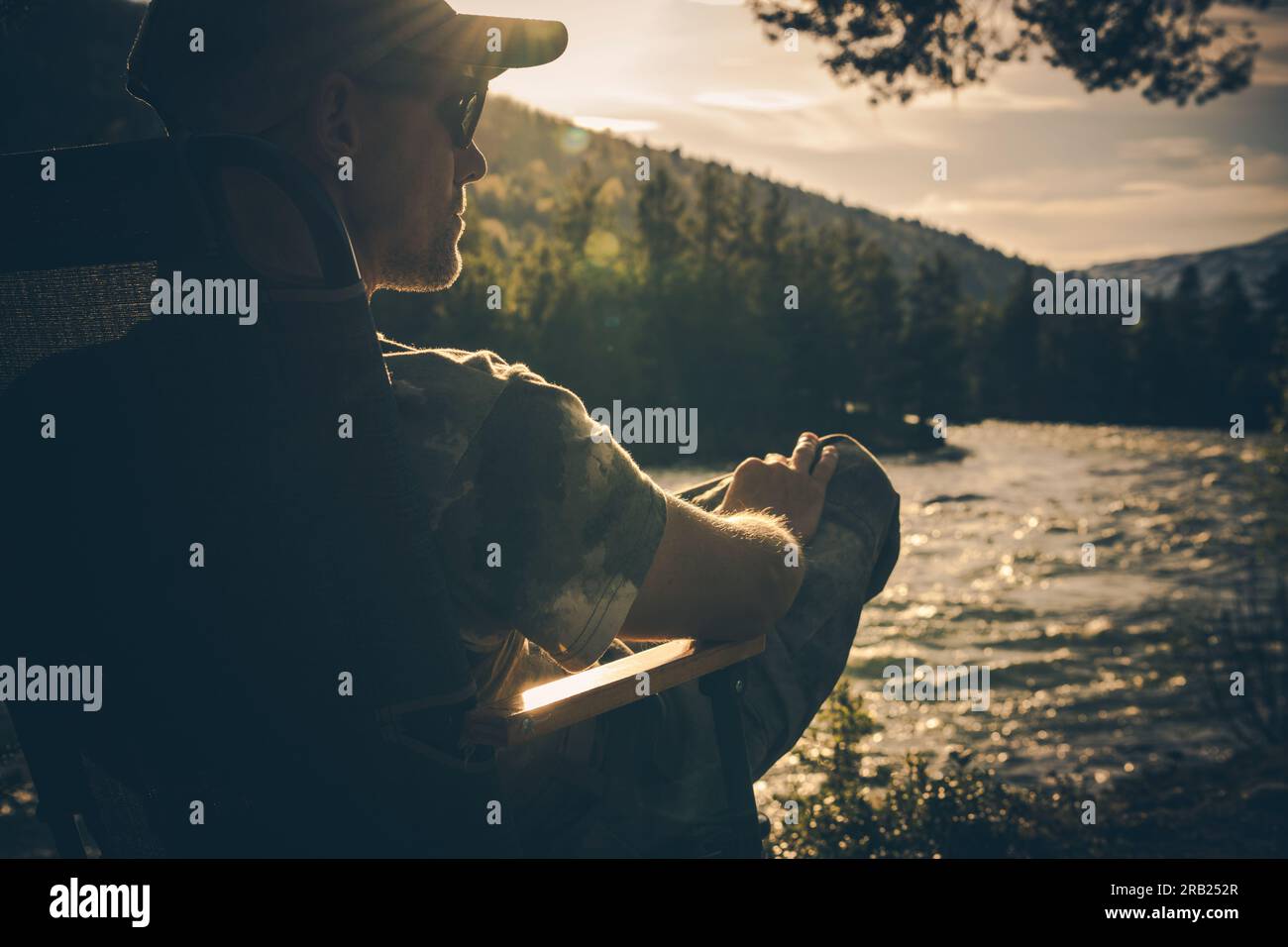Rückansicht eines entspannten Mannes, der im Campingstuhl am Fluss sitzt und den Sonnenschein und die wunderschöne Tierwelt genießt. Stockfoto