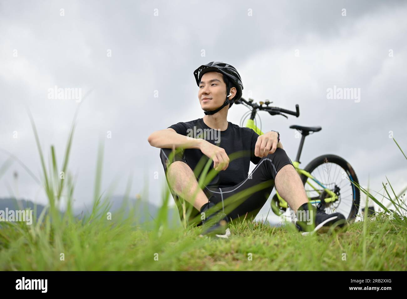 Ein glücklicher junger asiatischer Mann in Sportbekleidung ruht nach einer langen Radtour in den Bergen auf dem Gras. Männlicher Radfahrer, gesunder Lebensstil Stockfoto