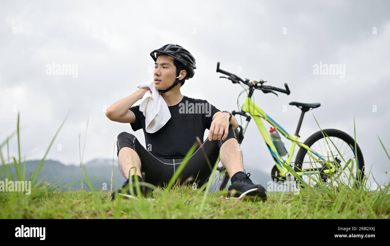 Ein müder junger asiatischer Mann in Sportbekleidung ruht nach einer langen Radtour in den Bergen auf dem Gras. Stockfoto