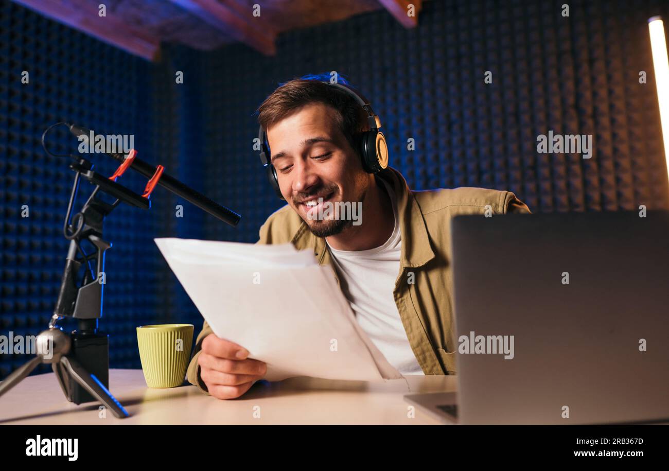 Lächelnder Radio-Moderator mit Kopfhörern, der Nachrichten aus Papier in das Studio-Mikrofon eines Radiosenders mit Neonlichtern liest. Ich schaue in die Kamera Stockfoto