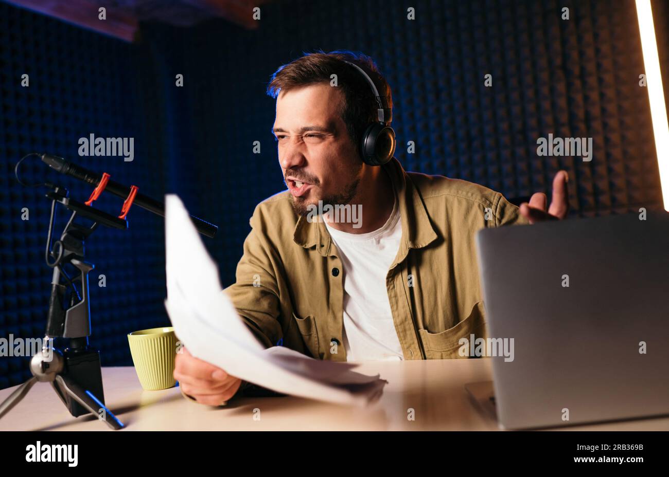 Radio-Moderator mit Kopfhörern, der Nachrichten aus Papier in das Studio-Mikrofon eines Radiosenders mit Neonlichtern liest Stockfoto