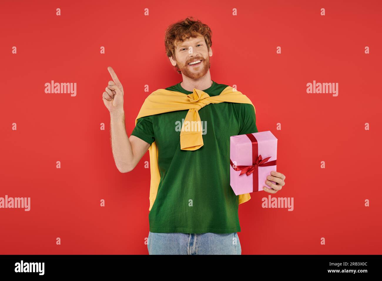 Feier, fröhlicher rothaariger Mann mit Bart in lässiger Kleidung auf Korallenhintergrund, mit Geschenkbox, festlichen Anlässen, Geschenk, Mode und Trend Stockfoto