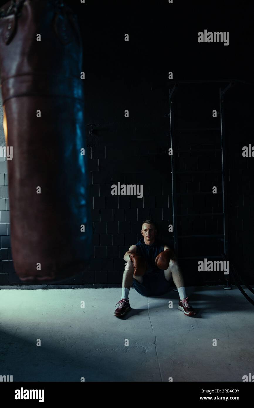 Müder Boxer in Boxhandschuhen, der nach dem Training im dunklen Fitnessstudio ruht. Stockfoto