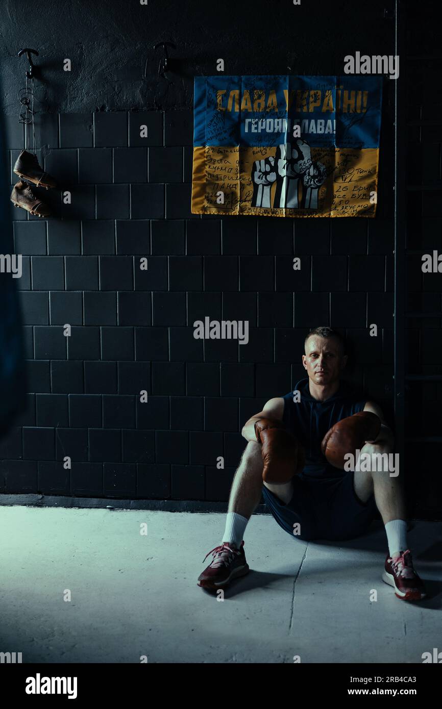 Müde Boxerin in Boxhandschuhen, die sich im dunklen Fitnessstudio ausruht, nachdem sie vor dem Hintergrund der ukrainischen Flagge trainiert hat, mit Unterschriften von ukrainischen Kriegern und Inscr Stockfoto