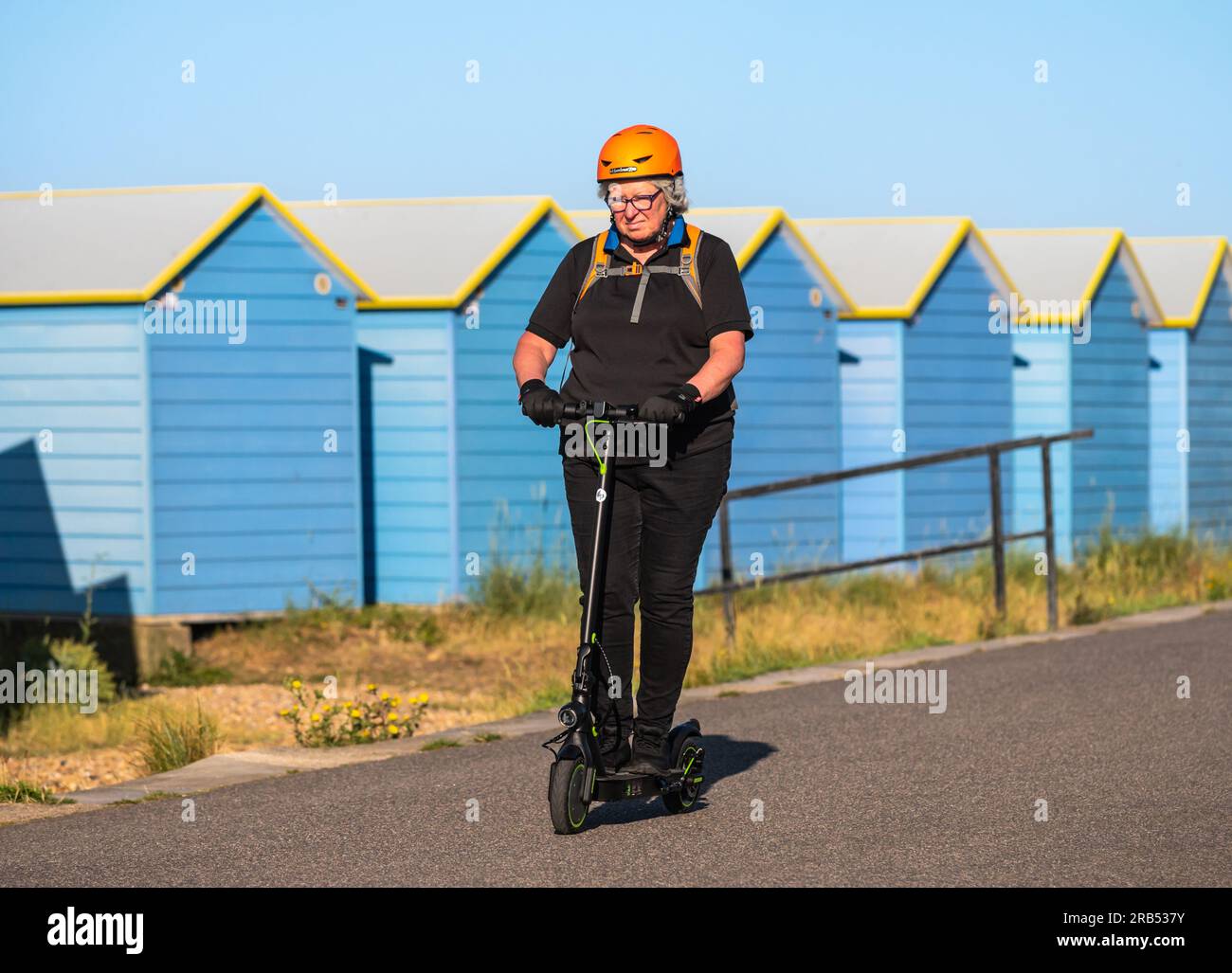 Seniorin mit Helm, die an einem warmen, sonnigen Tag in Großbritannien auf einem Elektroroller oder E-Scooter entlang einer Strandpromenade fährt. Stockfoto