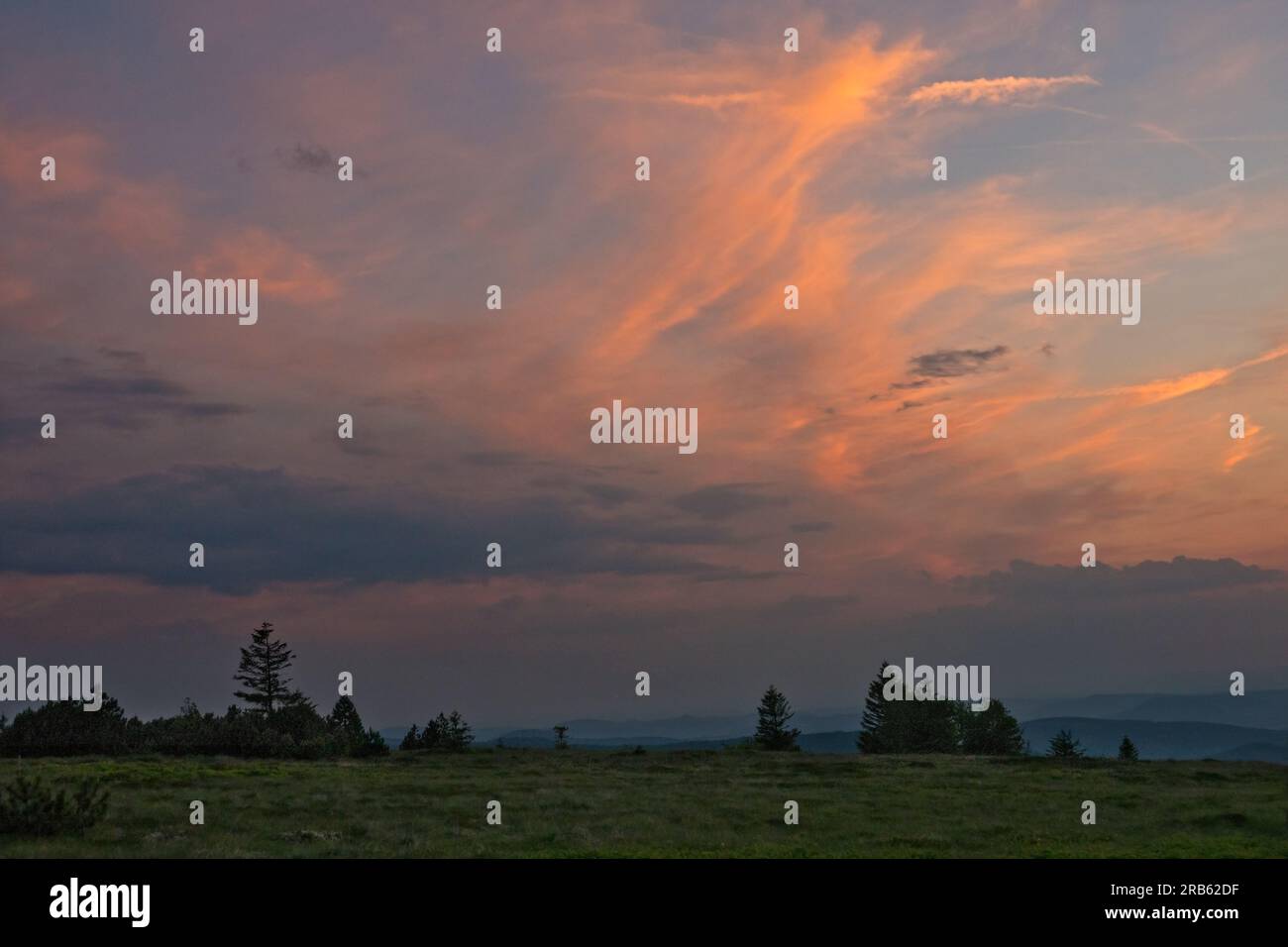 Zirrus-Wolke, orange beim letzten Licht der untergehenden Sonne, über einer hügeligen Landschaft in den Vogesen Stockfoto