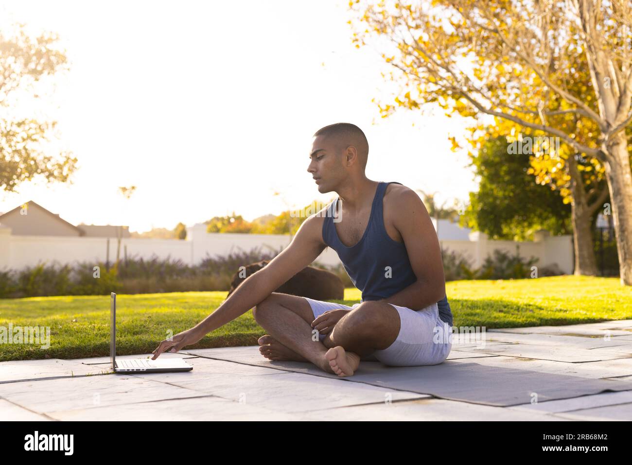 Fokussierter, birassistischer Mann, der Yoga praktiziert, sitzt im sonnigen Garten mit einem Laptop. Sommer, Wohlbefinden, Kommunikation, Fitness und gesunder Lebensstil, unverändert. Stockfoto