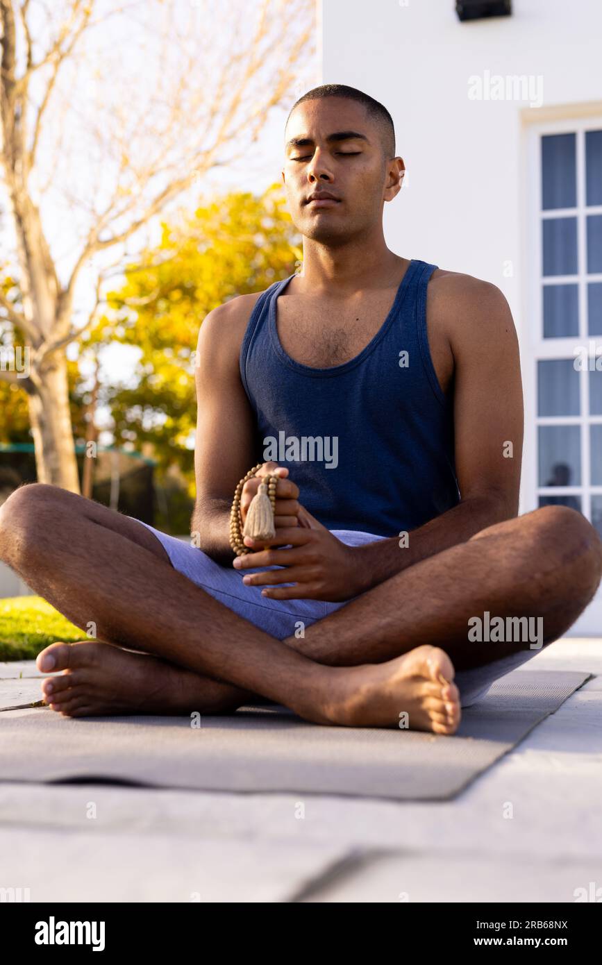 Ein ruhiger, birassistischer Mann, der Yoga-Meditation praktiziert und im sonnigen Garten mit geschlossenen Augen sitzt. Sommer, Wohlbefinden und gesunder Lebensstil, unverändert. Stockfoto