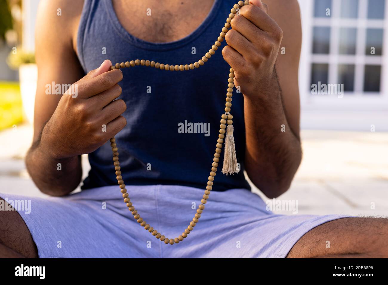 Mittelteil eines birassischen Mannes, der Yoga-Meditation praktiziert und im sonnigen Garten sitzt und Perlen hält. Mala Perlen, Sommer, Wohlbefinden, Fitness und gesundes Lifest Stockfoto