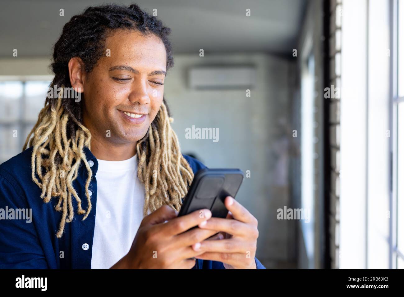 Glücklicher birassistischer, lässiger Geschäftsmann mit Dreadlocks, der in einem sonnigen Büro ein Smartphone am Fenster benutzt. Arbeit, Arbeit, Kommunikation und ungezwungenes Büro, unveränderlich Stockfoto