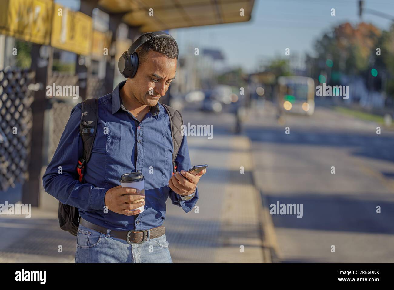 Ein junger latino mit Kopfhörern, der an der Bushaltestelle auf sein Handy schaut. Stockfoto