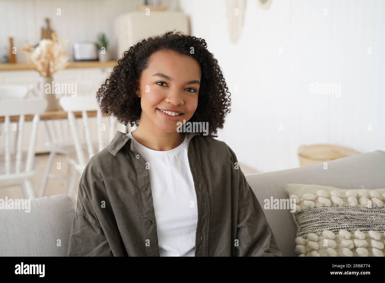 Ein glückliches birassisches Mädchen mit Afro-Haaren sitzt auf dem Sofa, lächelt im gemütlichen Wohnzimmer, schaut in die Kamera. Stockfoto