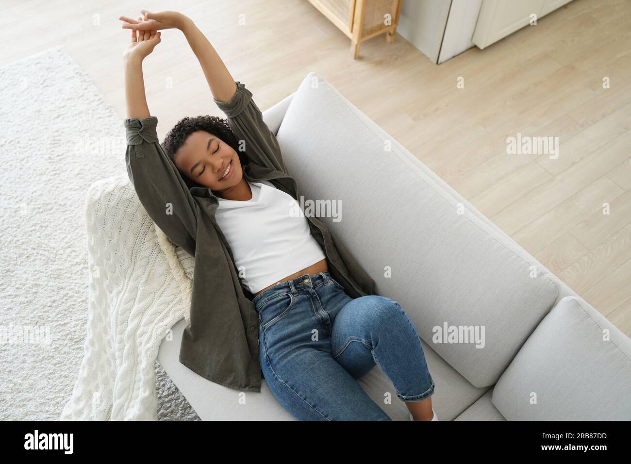 Ein lächelndes Mädchen dehnt sich nach einem Nickerchen auf dem bequemen Sofa zu Hause. Glücklicher Teenager entspannt sich und genießt einen ruhigen Wochenendtrip. Stockfoto