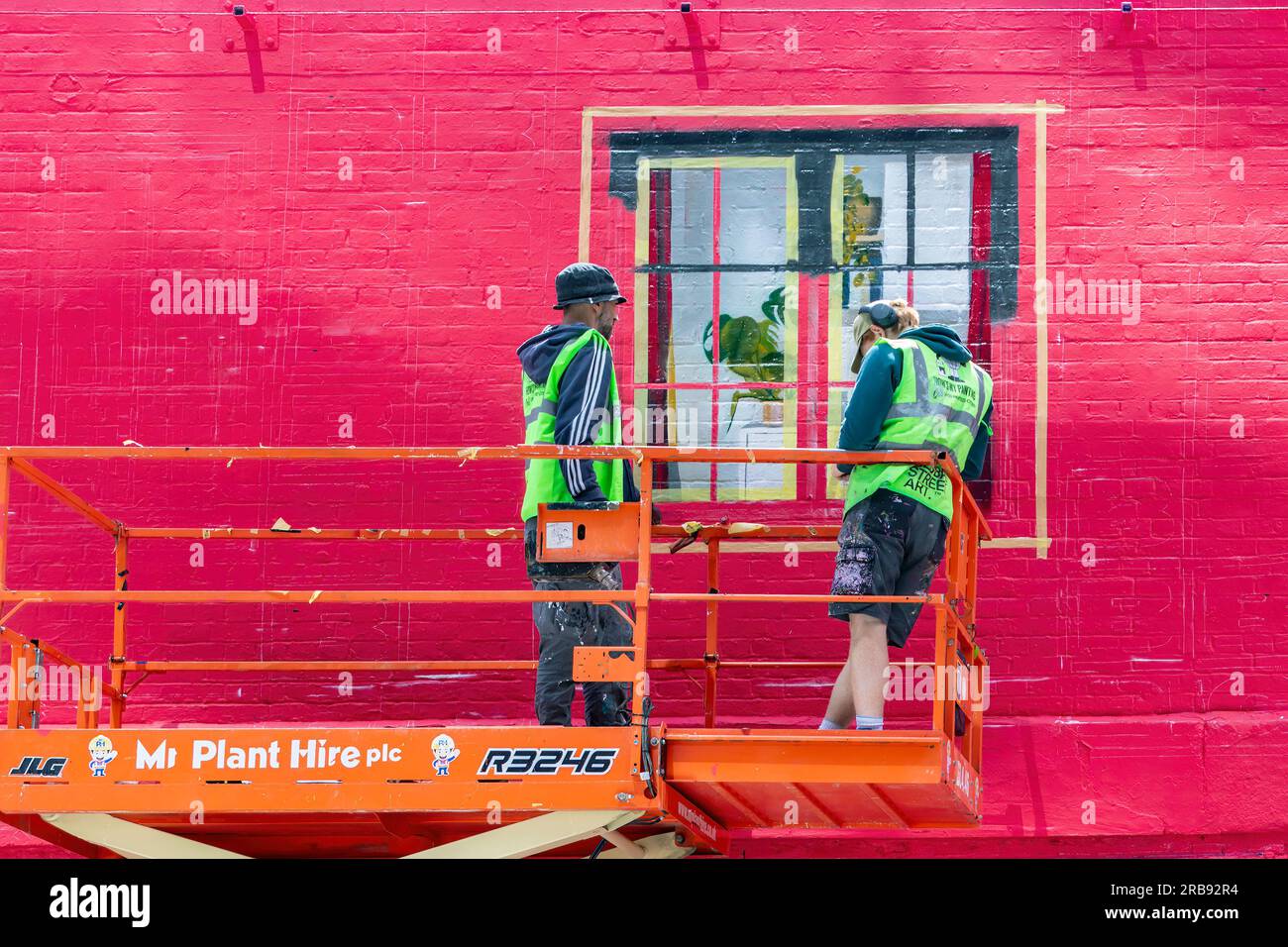 Zwei Arbeiter in leuchtend leuchtenden Jacken auf hoher Plattform, an einer Wandmalerei Kunst eines Fensters auf einer roten Ziegelwand am Gebäude shoreditch london uk Stockfoto