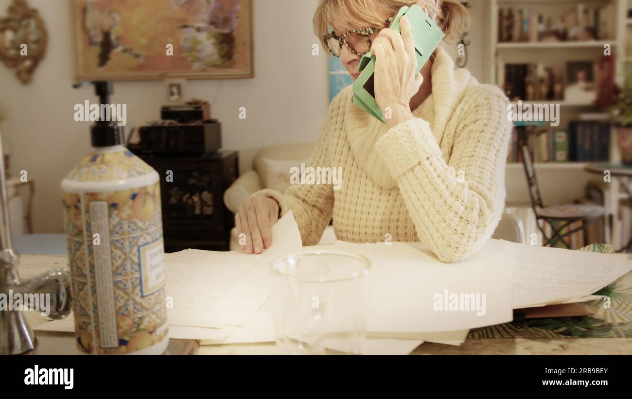 Auf dem Küchentisch legt eine Frau Papiere und Dokumente aus, während sie am Telefon erklärt oder fragt. Stockfoto