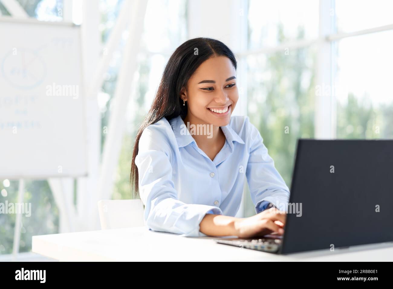 Lächelnde afroamerikanische Geschäftsfrau, die auf einem Laptop am Schreibtisch im modernen Büro sitzt. Geschäftserfolg Stockfoto