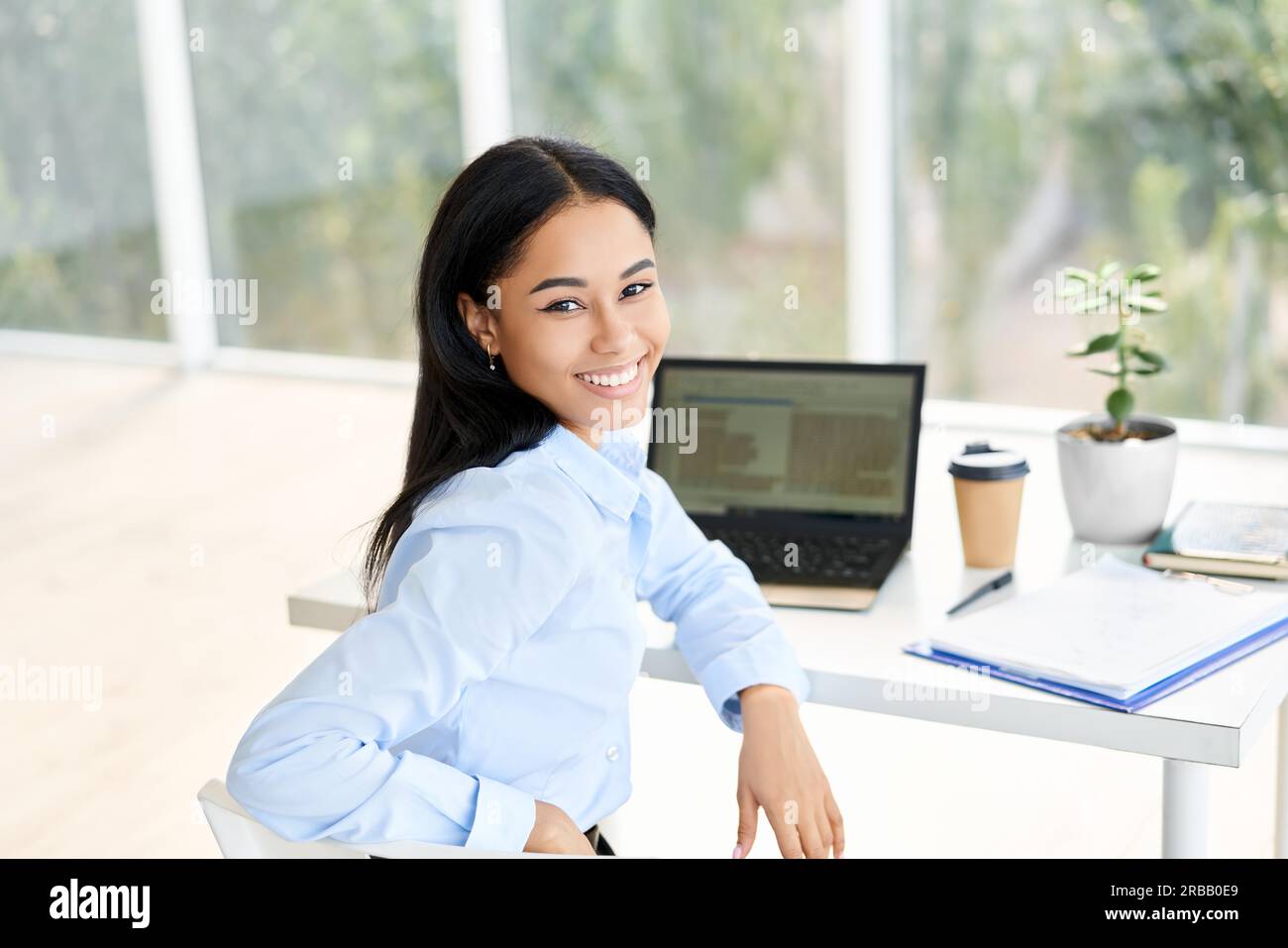 Lächelnde junge afroamerikanische Geschäftsfrau posiert an ihrem Schreibtisch in einem hellen modernen Büro. Business Success Concept Stockfoto