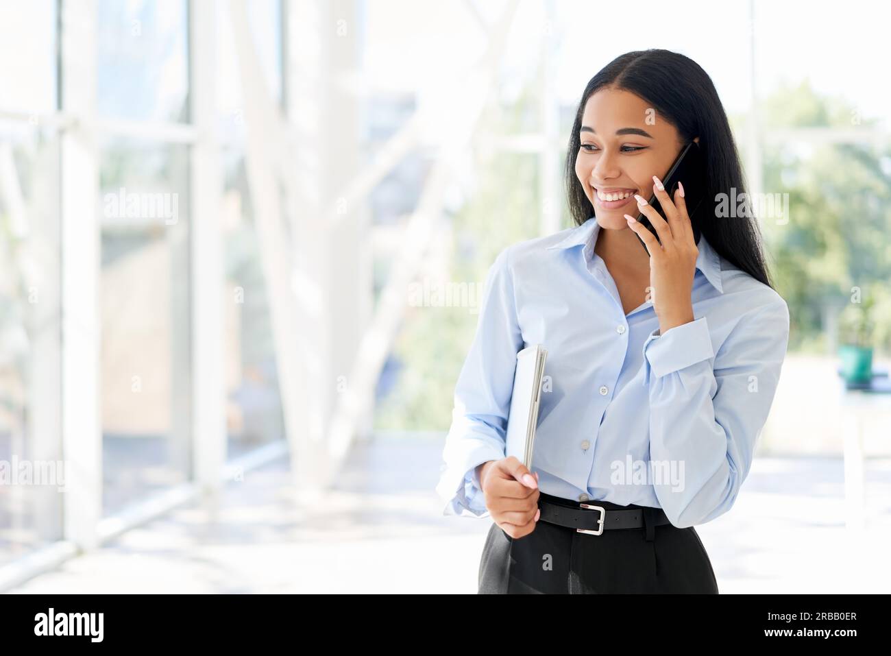 Lächelnde afroamerikanische Geschäftsfrau telefoniert in einem offenen, modernen Büro mit Panoramafenstern im Hintergrund. Speicherplatz kopieren. Kommunikation. Stockfoto