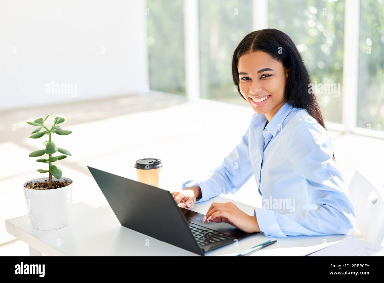 Lächelnde afroamerikanische Geschäftsfrau sitzt an ihrem Schreibtisch und arbeitet an einem Laptop in einem modernen Büro Stockfoto