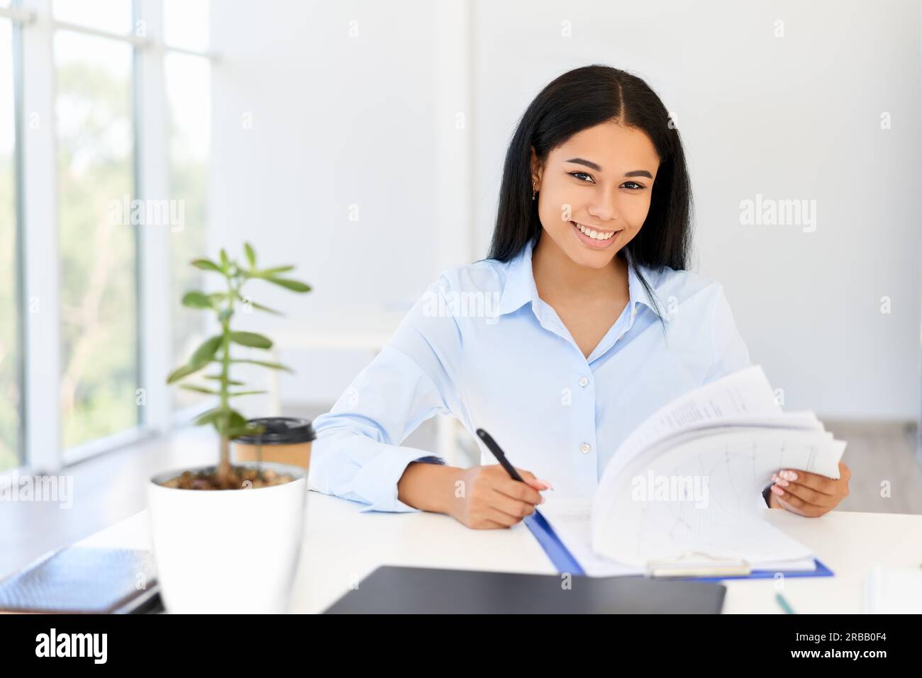 Lächelnde junge afroamerikanische Geschäftsfrau, die in einem hellen, modernen Büro auf ihrem Schreibtisch arbeitet. Frau im Geschäft, Erfolgskonzept Stockfoto