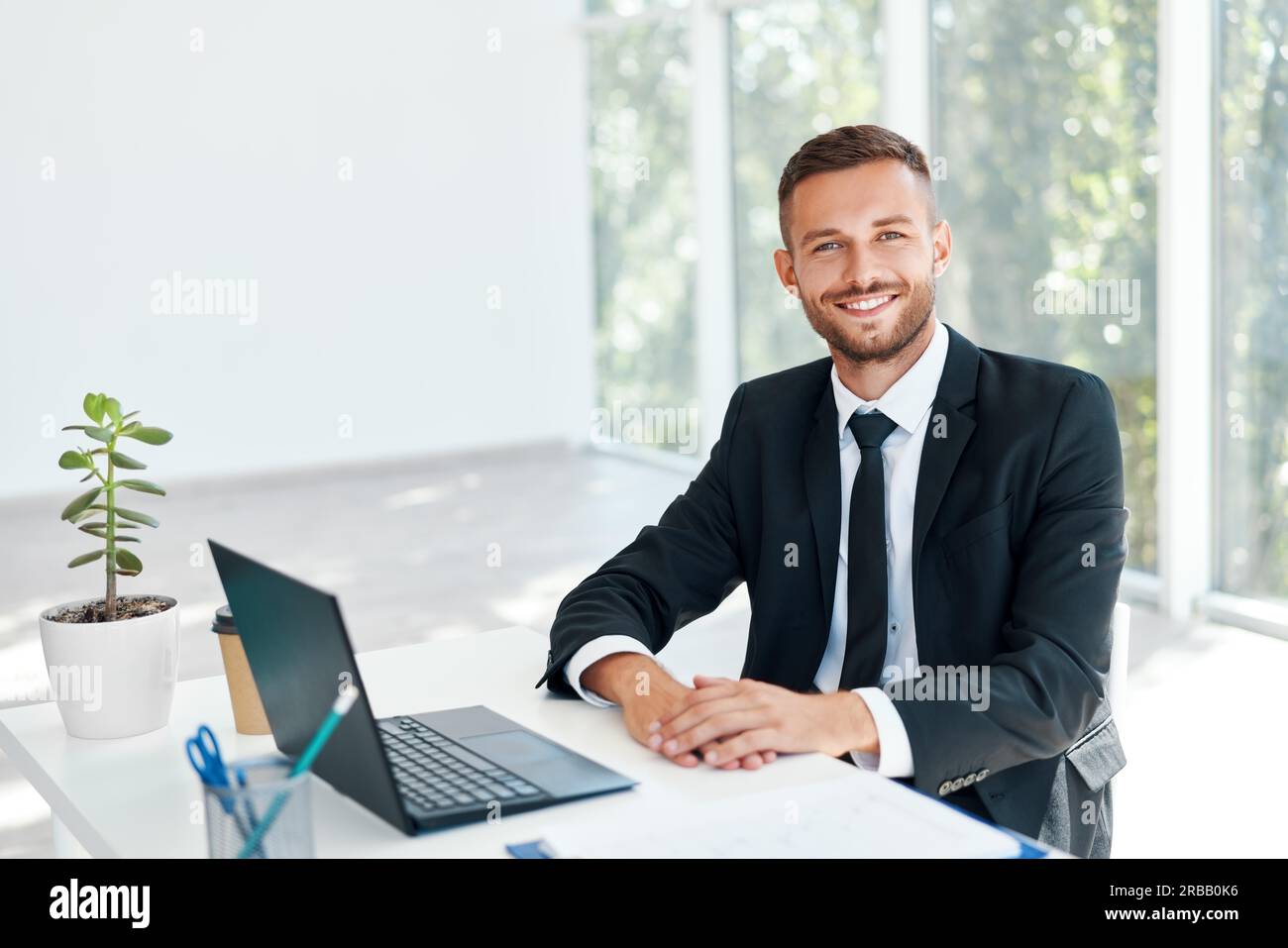 Stilvoll lächelnder Geschäftsmann im eleganten Anzug sitzt an seinem Schreibtisch in einem hellen modernen Büro. Business Success Concept Stockfoto