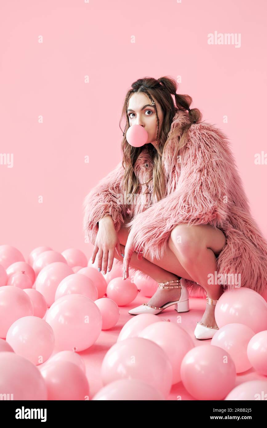 Lustige Hipster Frau Blasen Blase mit Kaugummi sitzen in vielen rosa Luftballons Hintergrund Stockfoto