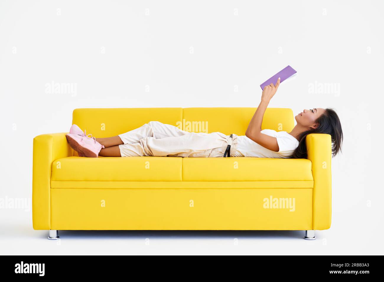 Hübsche asiatische Frau, die Buch liest, liegt auf der gemütlichen gelben Couch, Konzepte von Zuhause und Komfort, Hobby, Freizeit Stockfoto