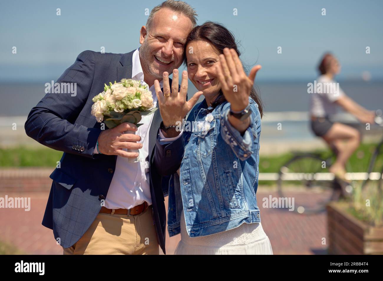 Glücklich romantisch lachend frisch vermähltes Paar, das ihre Ringe zeigt, während sie zusammen am Meer posieren, wobei der Mann den Brautstrauß in einem hält Stockfoto