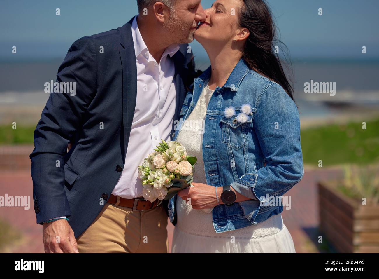 Glückliches, romantisches, reifes, frisch verheiratetes Paar an ihrem Hochzeitstag, küsst sich draußen auf einer Promenade vor dem Hintergrund des Ozeans und trägt formelle Kleidung und Stockfoto